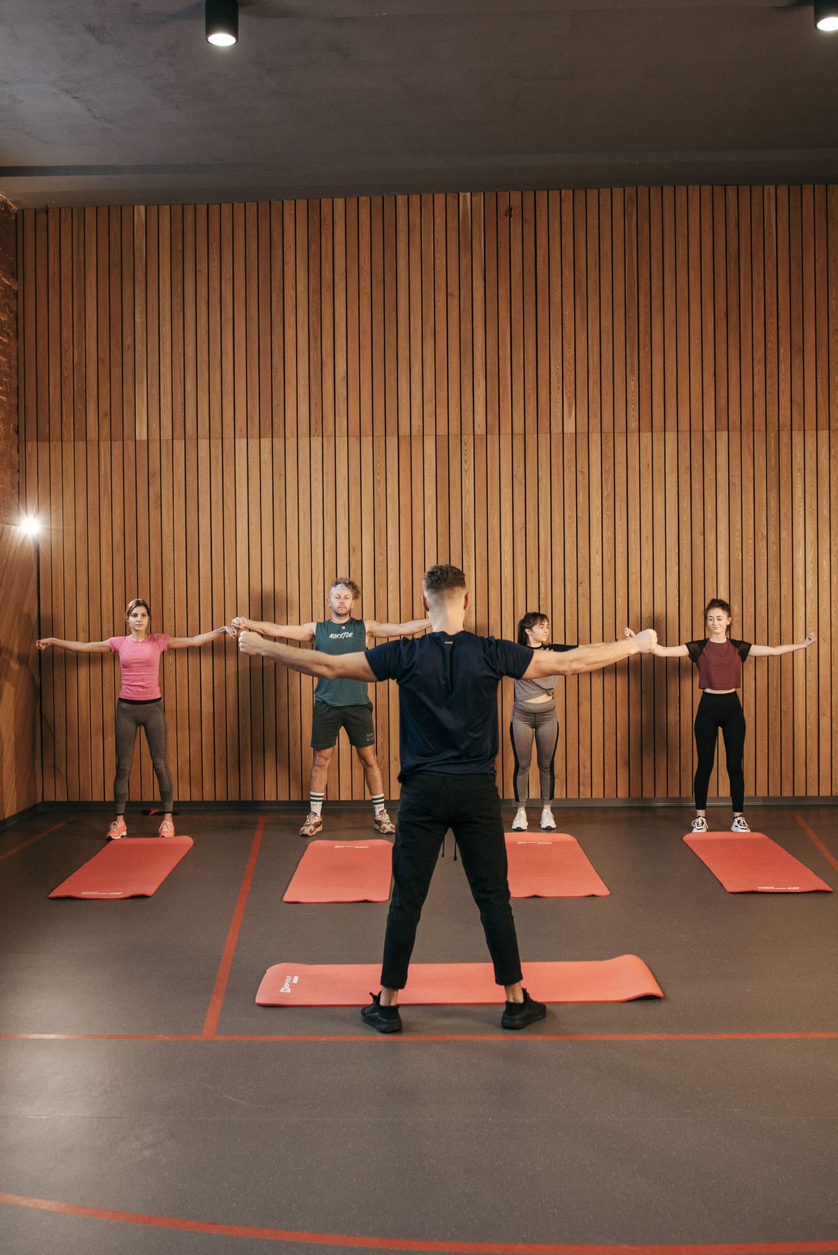 Four adults on mats in a studio, following the movement of a coach standing in front of them.
