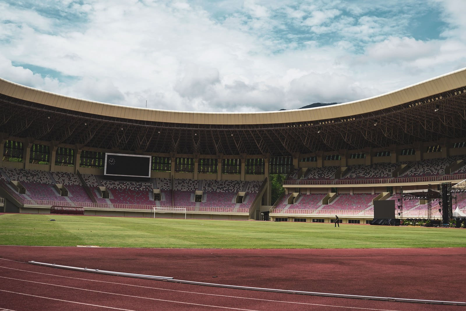 An empty athletics stadium seen from the track, the lanes and the infield under a wide clouded sky.