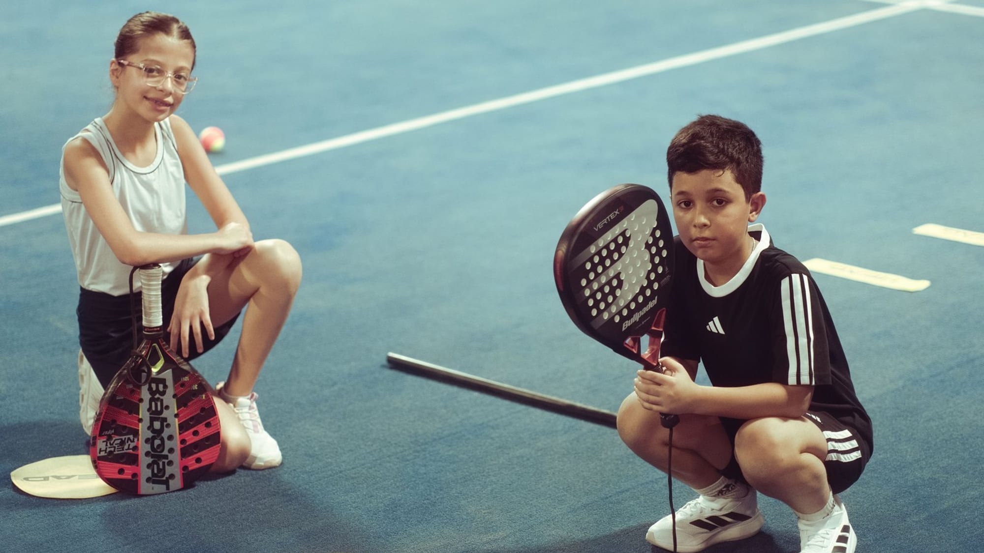 Two children crouching on a blue padel court, each holding a padel racket, with a training ball and floor markers beside them.