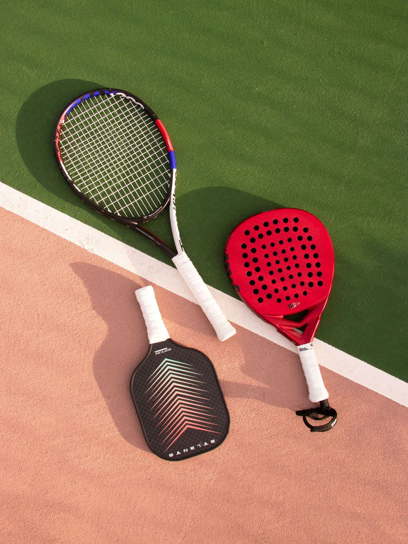 A tennis racket, a padel racket and a pickleball paddle laid out across the green turf and terracotta of a court, seen from above.