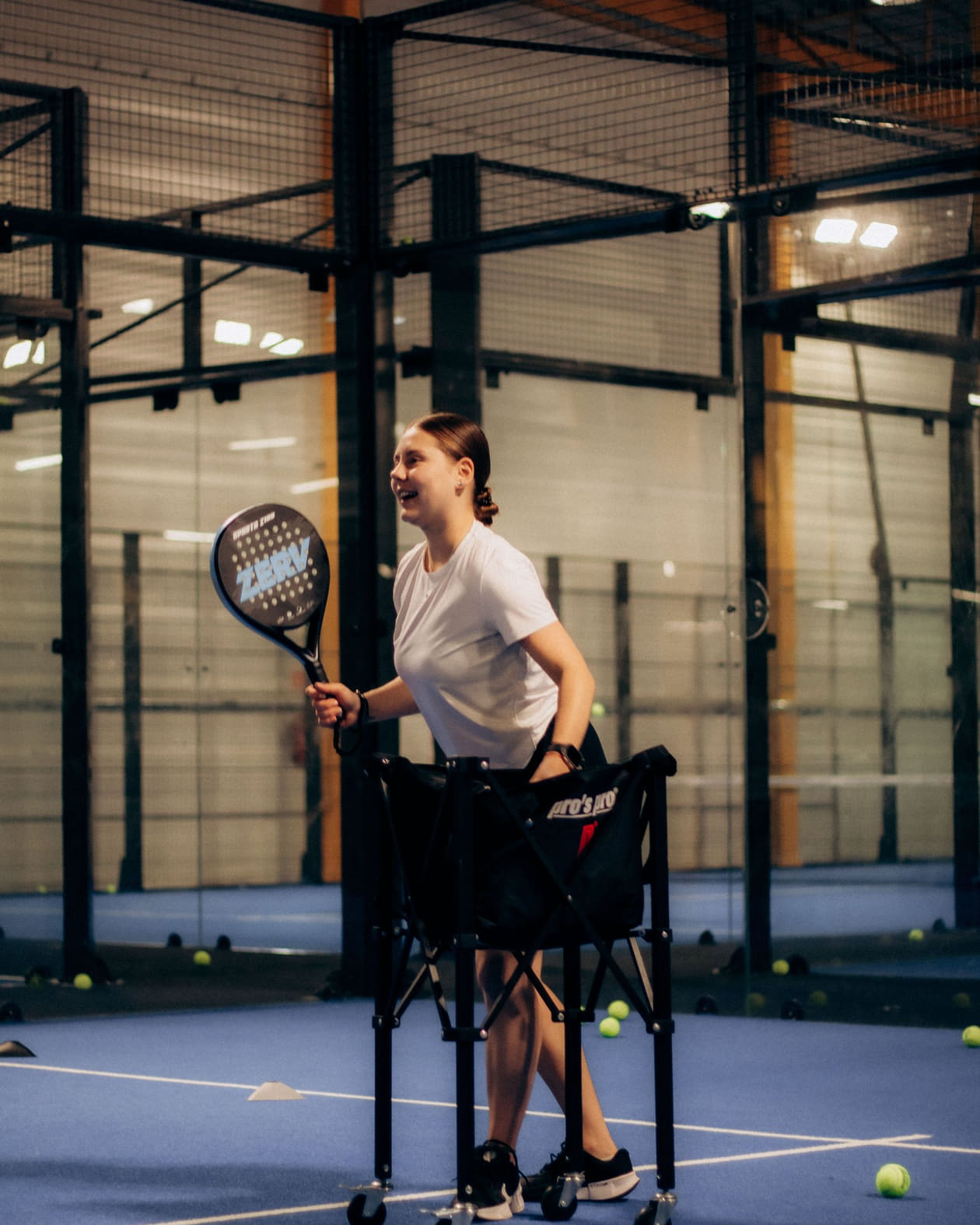 A person laughing on an indoor padel court, racket in hand, beside a cart of balls.