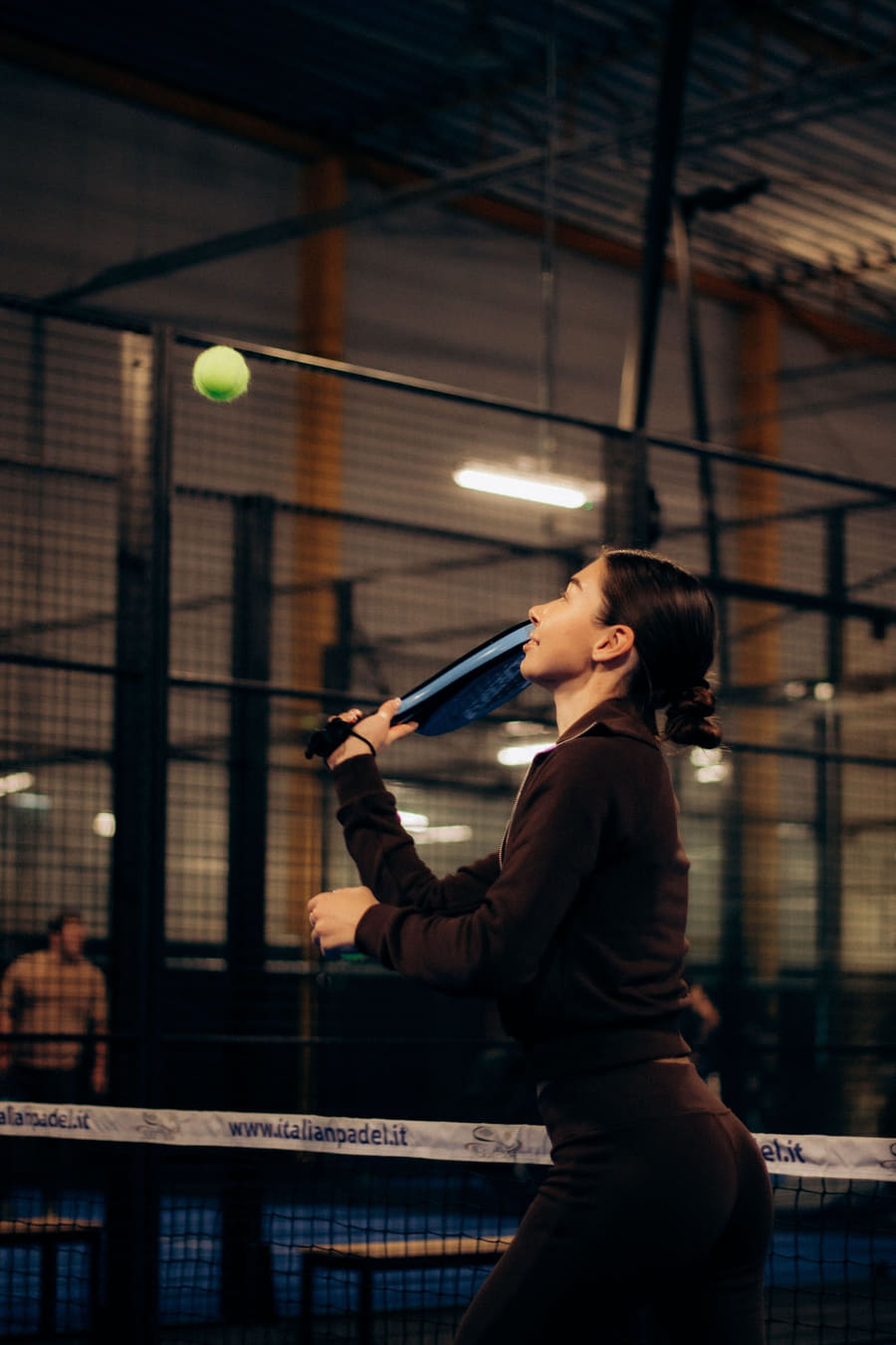 A player at the net on a dark indoor padel court, racket raised, the ball in the air above them.