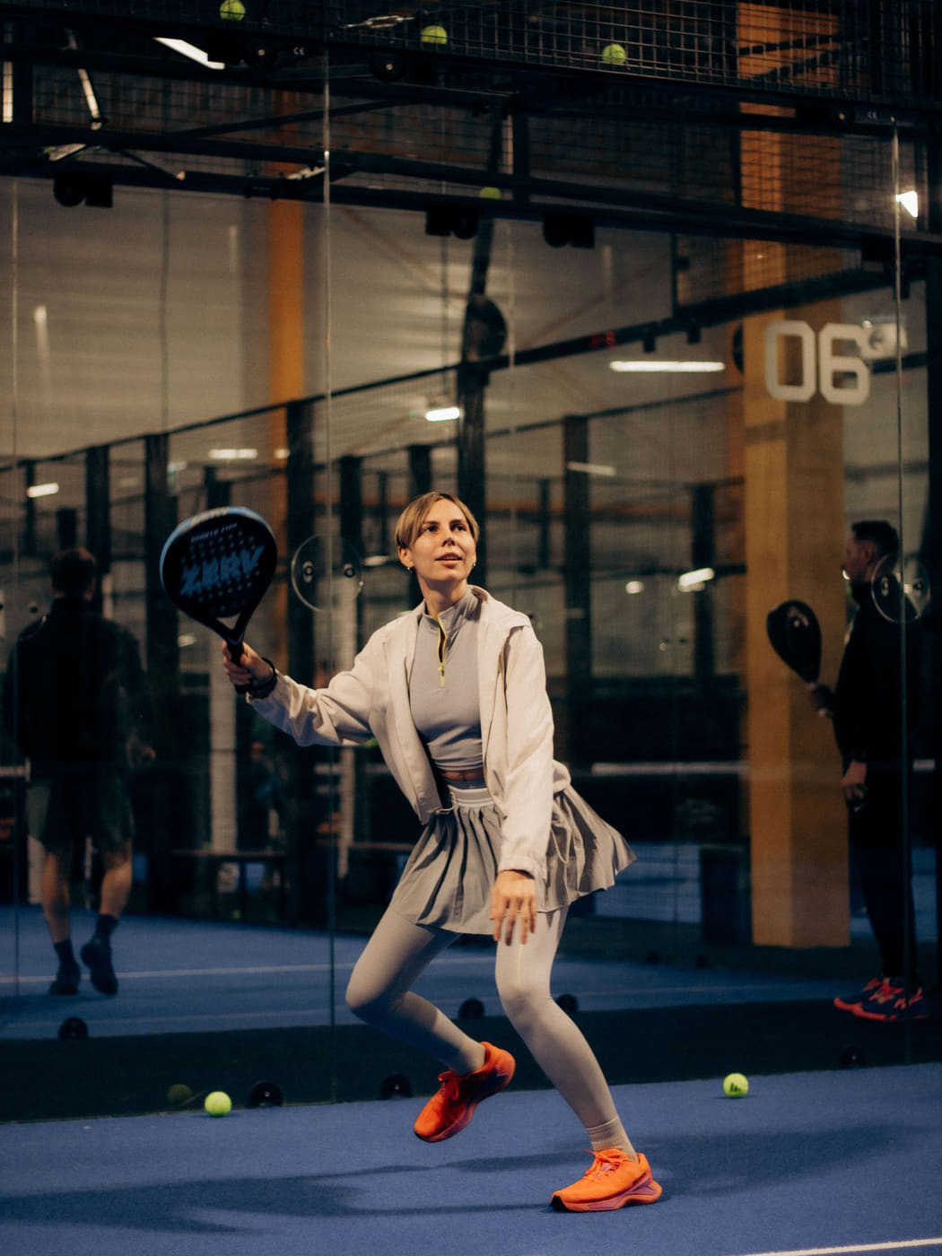 A player reaching wide for a forehand on an indoor padel court, the glass wall behind them.