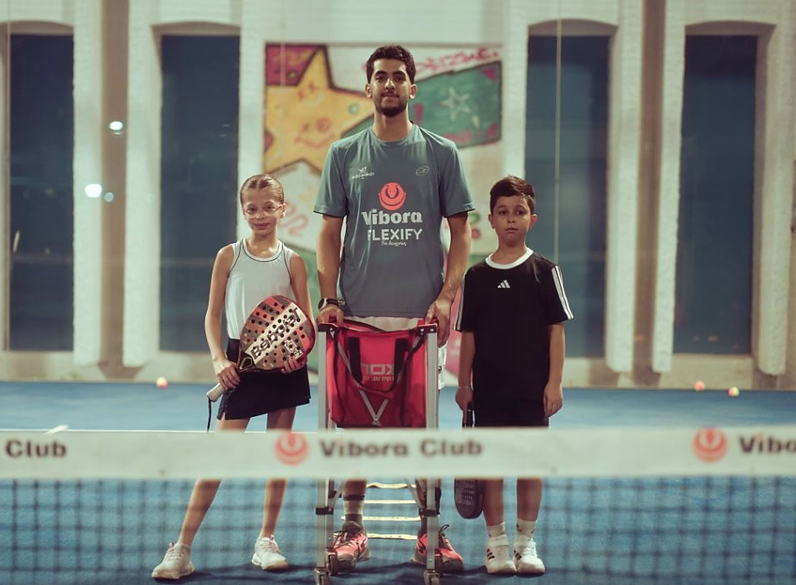 A coach standing at a padel net between two children holding rackets, a basket of balls in front of them.