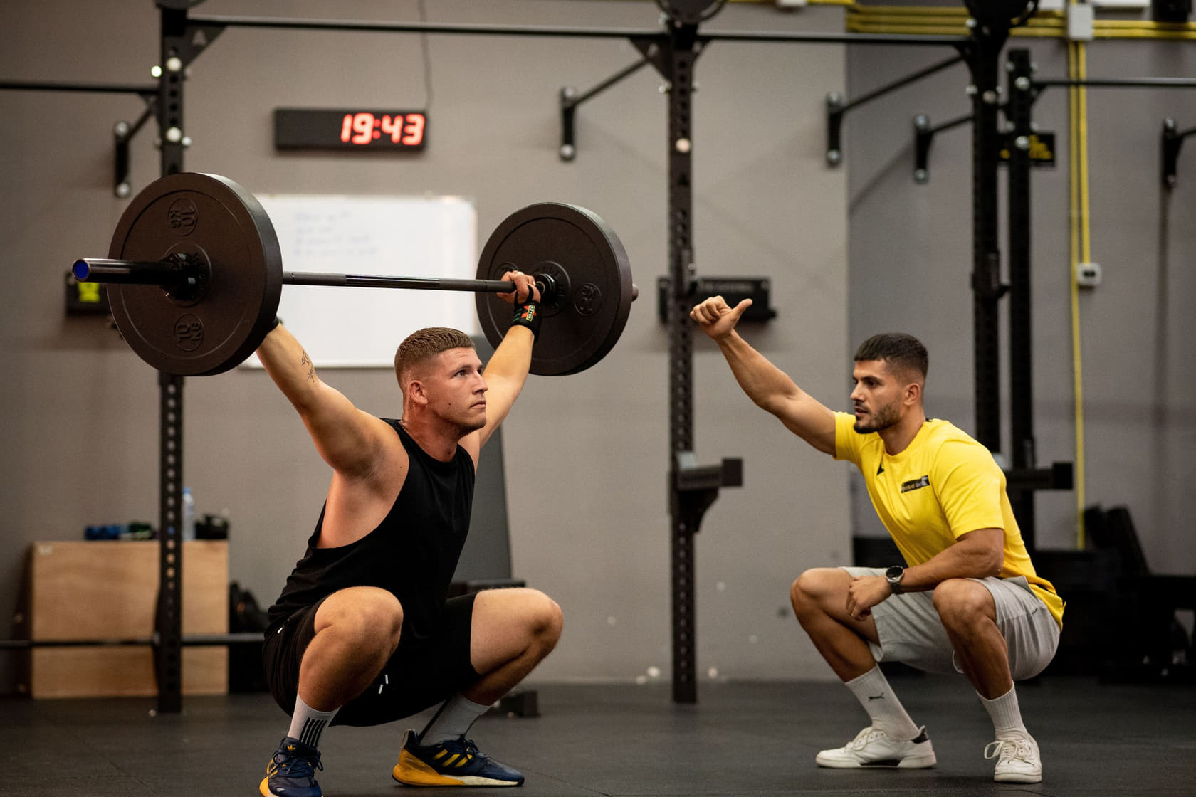 One person holding a barbell overhead in a deep squat while a second crouches beside them, giving a cue.