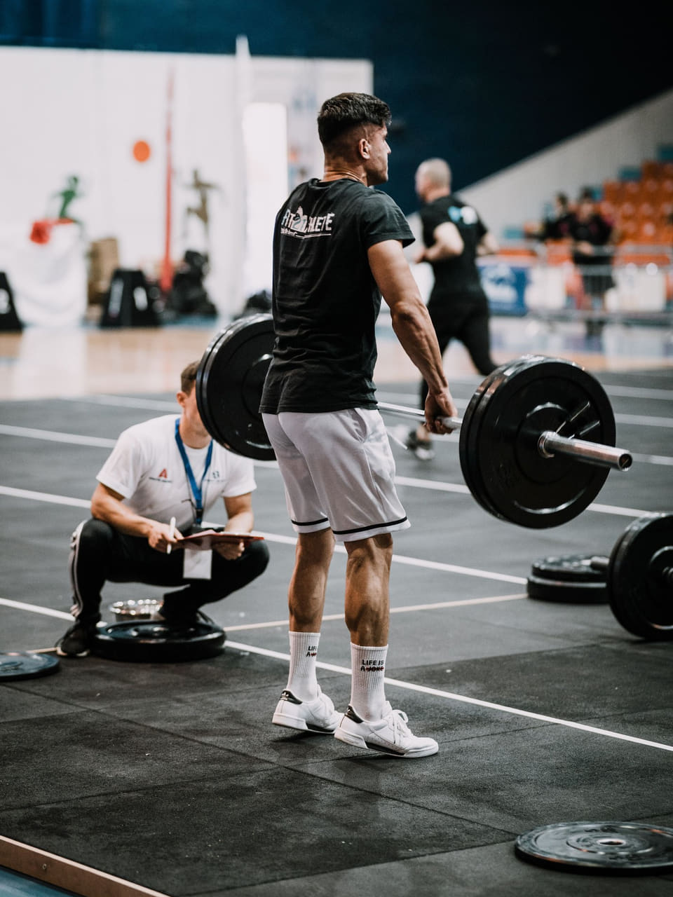 A lifter standing with a loaded barbell while a second person crouches behind them, writing on a clipboard.