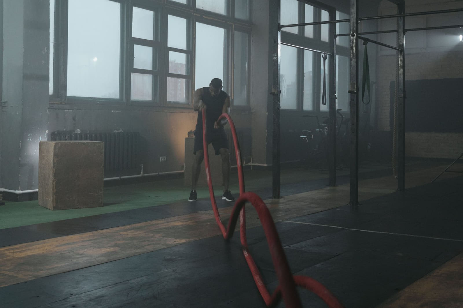 A single figure working battle ropes across the floor of a dark, hazy gym, tall windows behind.